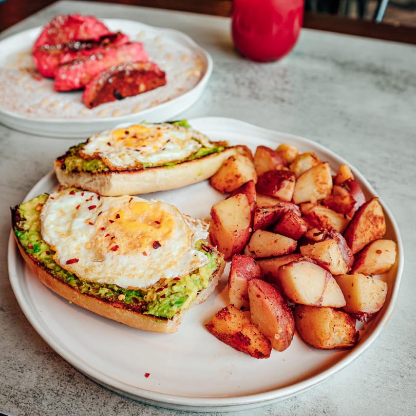Avocado toast with fried eggs and roasted potatoes, with pink French toast in the background.