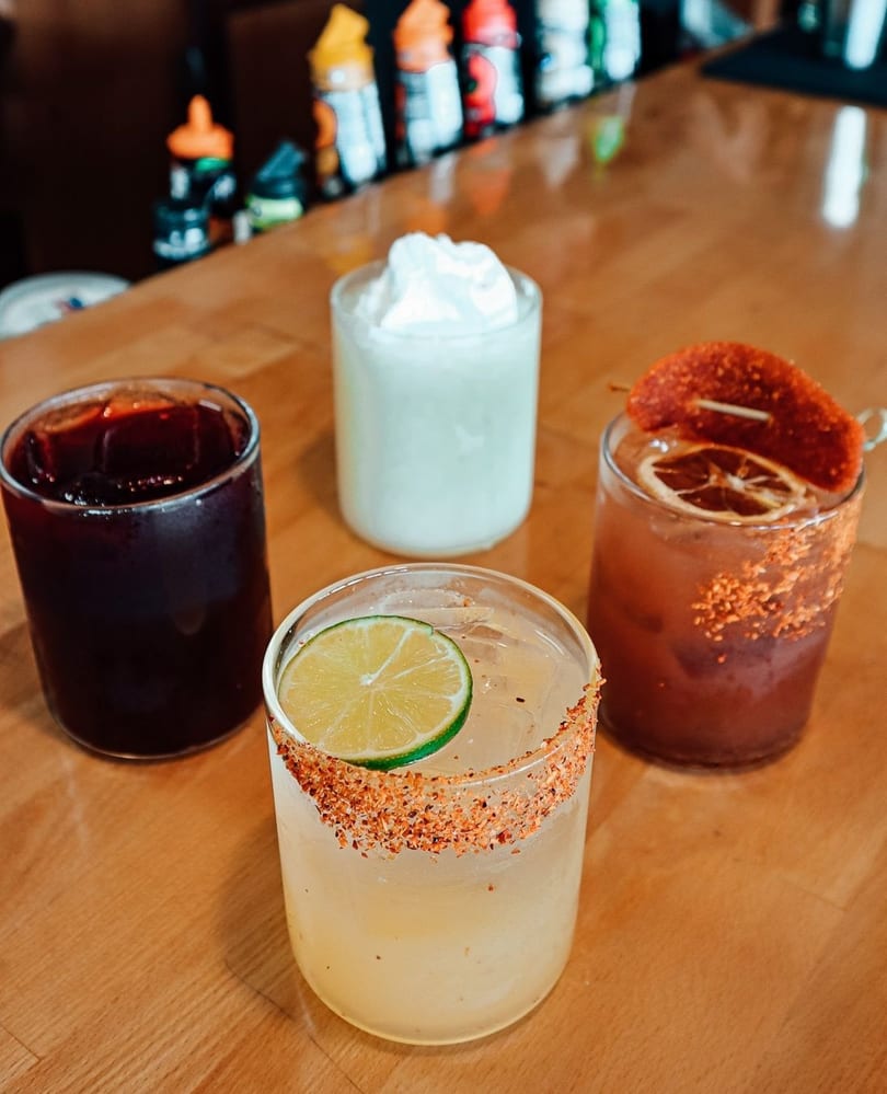 Four colorful cocktails, including a margarita with a lime, on a wooden bar.
